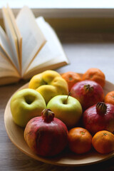 Wooden bowl filled with various seasonal fruit and open book on a table. Selective focus.