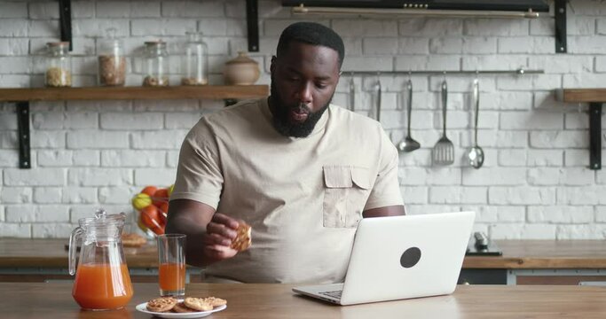 African male freelancer eats cookies and works at the computer while sitting at the kitchen table at home.