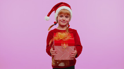 Funny toddler girl wears red New Year sweater and hat presenting Christmas gift box stretches out hands isolated on violet studio wall background. Happy Christmas celebration holiday shopping sale eve