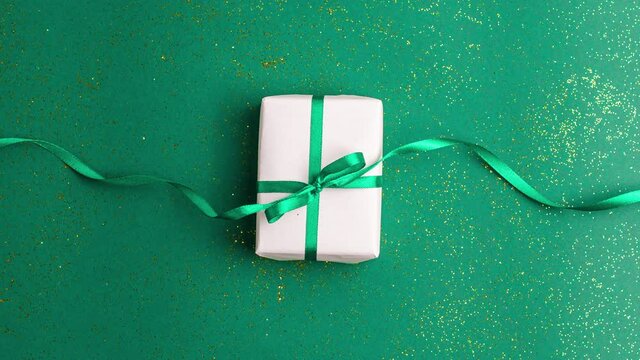 White gift box with a emerald green satin ribbon bow on green background with gold colored glitter. Stop motion animation Christmas Holidays and present concept flat lay with copy space.