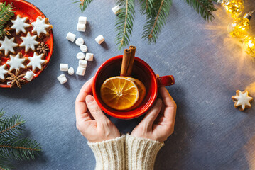 Woman holding in hands hot spicy Christmas tea with cinnamon and oranges, cookies and marshmallow on blue blanket. Xmas concept, top view.