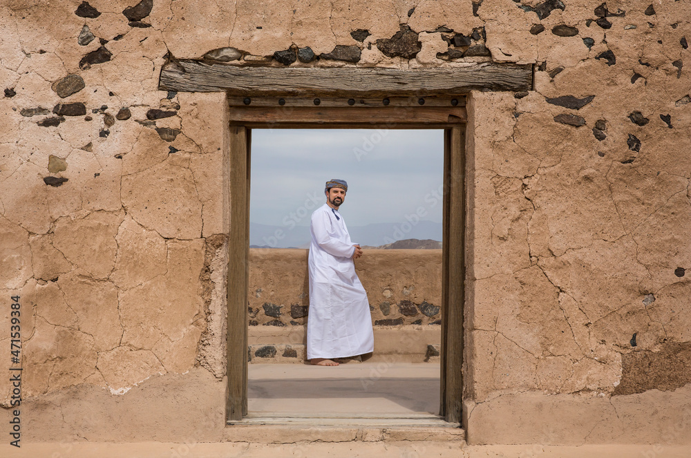 Poster omani man in traditional outfit at a doorway of an old house