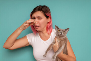 Young beautiful woman standing against blue background, holding her nose because of a bad smell coming out from the cat. - obrazy, fototapety, plakaty