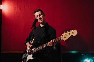 Obraz premium Talented handsome young guitarist man singing a song in studio recording on red background surrounded by instruments. Passion, hobby, singer, electric guitar
