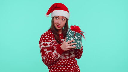 Funny adult girl 20s years old wears red New Year sweater deer antlers holding gift, received present and interested in what inside box on blue background studio. Happy Christmas holiday celebration