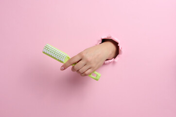 female hand holds a green plastic brush on a pink background. A part of the body sticks out of a hole with torn edges in a paper background