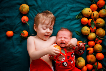 Fototapeta na wymiar The older brother wants to feed the Christmas newborn baby a tangerine, the baby cries. Children lie on a green blanket next to tangerines. High quality photo