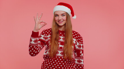 Adult girl in Christmas sweater looking approvingly at camera showing ok gesture, like sign positive something good. Young woman isolated on pink background. Happy New Year celebration merry holiday