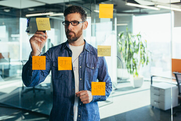 focused and thinking businessman writing on colorful notes attached to a glass wall, the...