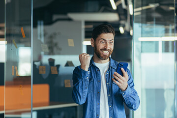 Excited happy business man with a beard, a professional winner, celebrating success by reading good news on a smartphone, feeling excited about mobile messaging