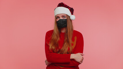 Woman in Christmas red sweater and hat wearing sterile face mask ppe to safe from coronavirus on lockdown quarantine. Adult girl indoors isolated on pink background. Covid-19 New Year celebration fail