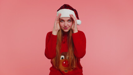 Woman in sweater Santa Christmas hat fooling around having closing eyes with hand and spying through, hiding and peeping isolated on red background studio. Happy New Year celebration merry holiday
