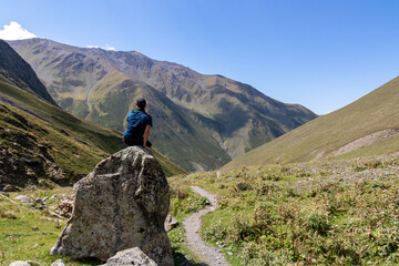 A male hiker sitting on a massive rock next to a hiking trail and enjoying the panoramic view on the high ridges of the mountain peaks in the Greater Caucasus Mountain Range in Georgia,Kazbegi Region.