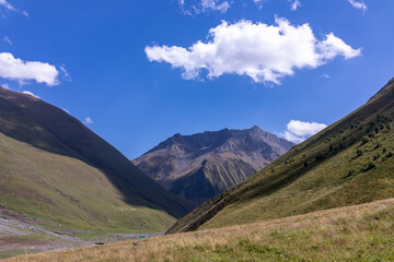 Fototapeta na wymiar A panoramic view on the high mountain peaks of the Chaukhi massif in the Greater Caucasus Mountain Range in Georgia, Kazbegi Region. A valley with lush green pastures. Wanderlust. Remote location.