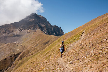 A woman on a hiking trail with a panoramic view on the Chaukhi Pass and its mountain peaks in the Greater Caucasus Mountain Range in Georgia, Kazbegi. The valley is lush green. Wanderlust.