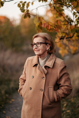 Beautiful and slim elderly woman in a woolen coat, scarf posing, standing under a beautiful tree with golden leaves in an autumn park, relaxing and enjoying the bright foliage, happy retirement