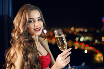 Adorable woman with long wavy hair in red dress drinking champagne during evening time. Blur background of city lights through panoramic windows. Winter holidays.