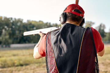 Rear view on caucasian Man in cap and headphones shooting at target on an outdoor shooting range at...