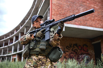 Fototapeta premium military female in gear with rifle with an optical sight. Female chase sport hunter in forest field with weapon near abandoned building, aiming rifle at target, focused or concentrated. alone