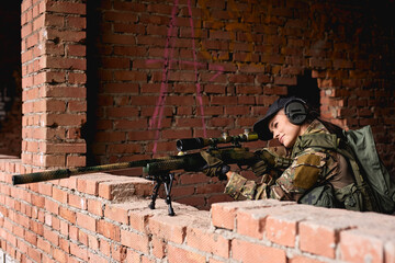 Fototapeta na wymiar caucasian female with sniper rifle with telescopic sight, keep defense in battle and aims at enemy. In Abandoned red brick wall building, female is looking at side. Side view on Soldier in action
