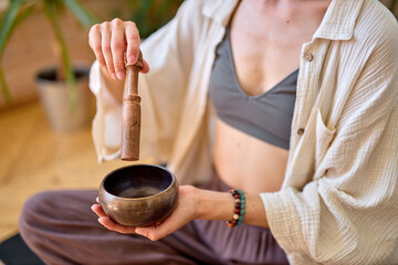 Fototapeta na wymiar Woman using singing bowl in sound healing therapy at home, sitting alone at home, unrecognizable female in domestic wear pajamas begins morning with yoga practice, sit relaxed. Close-up photo of hands