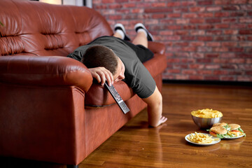 fat boy sleep on sofa in living room, young caucasian teen boy fall asleep while he was watching tv and eating crisps junk food, relaxed lazy boy is overeating unhealthy meal, at home
