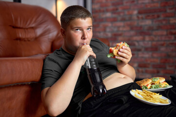 Overweight boy with fast food on floor near sofa at home, eating junk food drinking sweet beverages, lazy fat teenage boy in casual wear at home alone, having no control in eating behavior.
