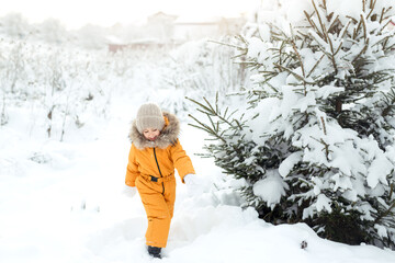 Fototapeta na wymiar A child in a yellow jumpsuit walks among fir trees in snowdrifts on a frosty cold day. The girl rejoices at the first December snowfall. Winter children's games and entertainment