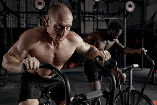 diverse sportsmen riding stationary bike while working out at gym, cross fit cardio training, shirtless black and caucasian men have intense training. healthy lifestyle. focus on caucasian male