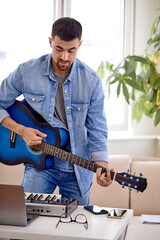 Man in denim jacket playing acoustic guitar, using laptop, performing music in studio, standing behind desk with piano, window in the background. Handsome caucasian male preparing for concert