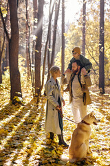 Fototapeta premium Da, mom and child boy walking with dog in warm sunny day in forest during leaf fall at autumn season. Lovely and carefree caucasian family consisted of parents, child and pet enjoy the walk