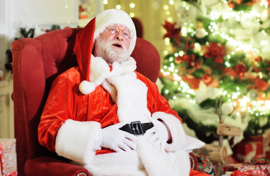 Santa Claus with a real gray beard in a traditional costume is sitting in an armchair and laughing holding his stomach.