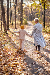 Fototapeta premium view from back on woman walking with son, having conversation, jumping and having fun. rear view. autumn season, sunny day. happy childhood, lifestyle, relationships, human emotions concept