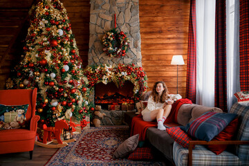 A beautiful girl is sitting on a sofa in a Christmas interior. Christmas tree, decorated fireplace, lots of gifts, Christmas theme. Cozy atmosphere.