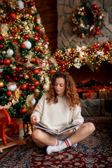 A beautiful girl is sitting on a sofa in a Christmas interior. Christmas tree, decorated fireplace, lots of gifts, Christmas theme. Cozy atmosphere. red-haired girl reading a book.