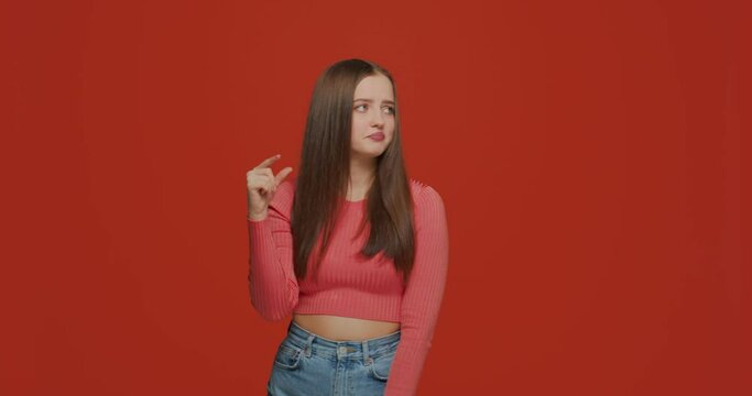 Young girl showing small size with fingers, sceptic about amount, gesturing on orange studio background