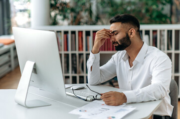 Naklejka premium Tired exhausted Indian man, company owner or office employee, sits at work desk with closed eyes, rubs his eyes, feeling headache, stress and fatigue from overtime work, need rest and break