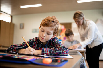 Portrait of smiling caucasian schoolboy writing test in his classroom and teacher in background.