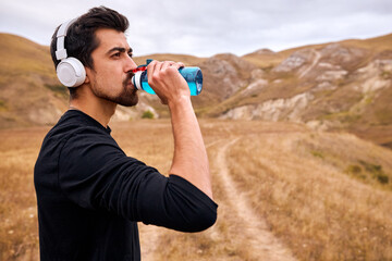 Sportsman take a sip of fresh water after jogging on fresh air in field among mountains, drinking, in headphones. Exhausted caucasian bearded guy in black sportswear having rest, taking a break