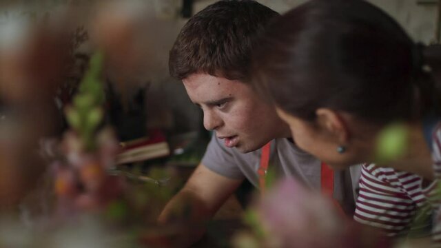Close up of young male florist with Down syndrome working on laptop with help of mentoring colleague indoors in flower shop.