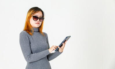 Fototapeta premium Studio shot of Asian young dyed hair female model in gray turtleneck sweater standing holding hand touching fashion black lens sunglasses holding tablet on white background