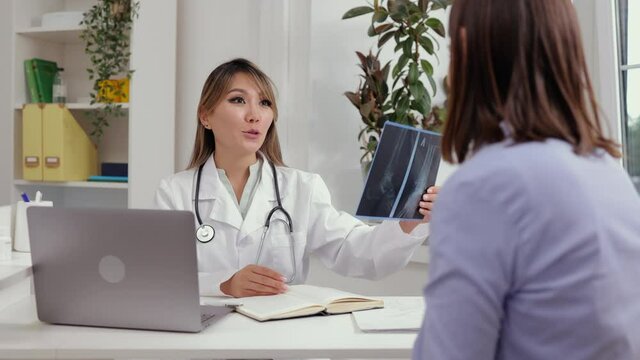 Doctor with patient at appointment. Positive Asian female therapist in white uniform shows x-ray picture to woman client visiting hospital office spbi 4k video