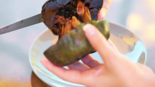 Macro closeup of female hands slicing in half one ripe soft diospyros nigra black sapote on white plate, chocolate pudding soapapple zapote prieto tropical fruit