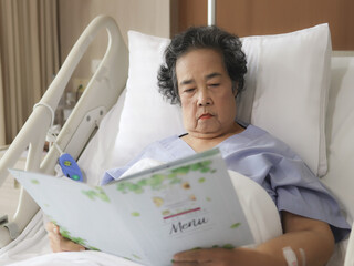 Asian senior female patient  sitting and reading food menu in hospital bed, ordering food. Elderly...