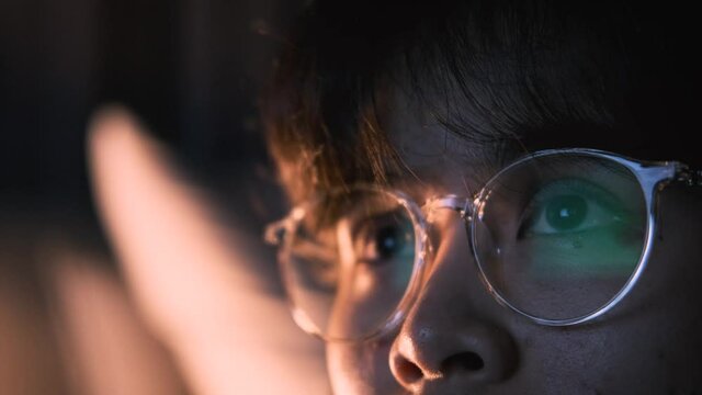 Close up view of focused women wears glasses for reducing eye strain blurred vision looking at television screen  with reflection using internet, watching, movie at nigh