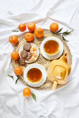 Two cups of tea, teapot, mandarins, chocolate cookies on the white bedsheet. Couple winter morning. Tea breakfast service. Cozy, warm atmosphere. Place for text.
