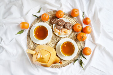 Two cups of tea, teapot, mandarins, chocolate cookies on the white bedsheet. Couple winter morning. Tea breakfast service. Cozy, warm atmosphere. Place for text.
