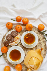 Two cups of tea, teapot, mandarins, chocolate cookies on the white bedsheet. Couple winter morning. Tea breakfast service. Cozy, warm atmosphere. Place for text.
