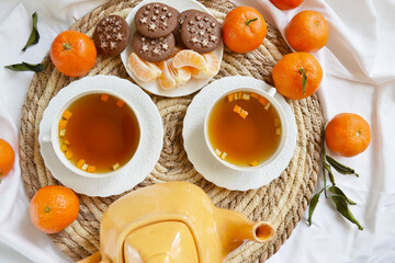 Two cups of tea, teapot, mandarins, chocolate cookies on the white bedsheet. Couple winter morning. Tea breakfast service. Cozy, warm atmosphere.