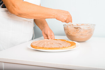 Close-up senior woman cook decorating layered cake with cream using spatula in kitchen indoors. Retired culinary hobby
